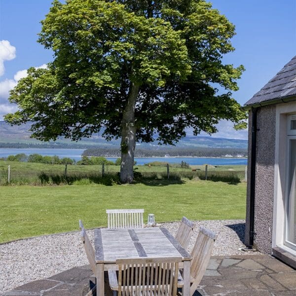 Outside seating area with table and chairs at Farthingworth holiday rental home, Skelbo, Dornoch with large tree in middle ground and view of Loch Fleet in background on sunny day