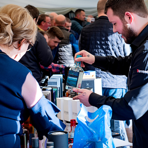 Depicts the Dornoch Whisky Festival Grand Tasting with members of the public trying drams at an exhibitors table.